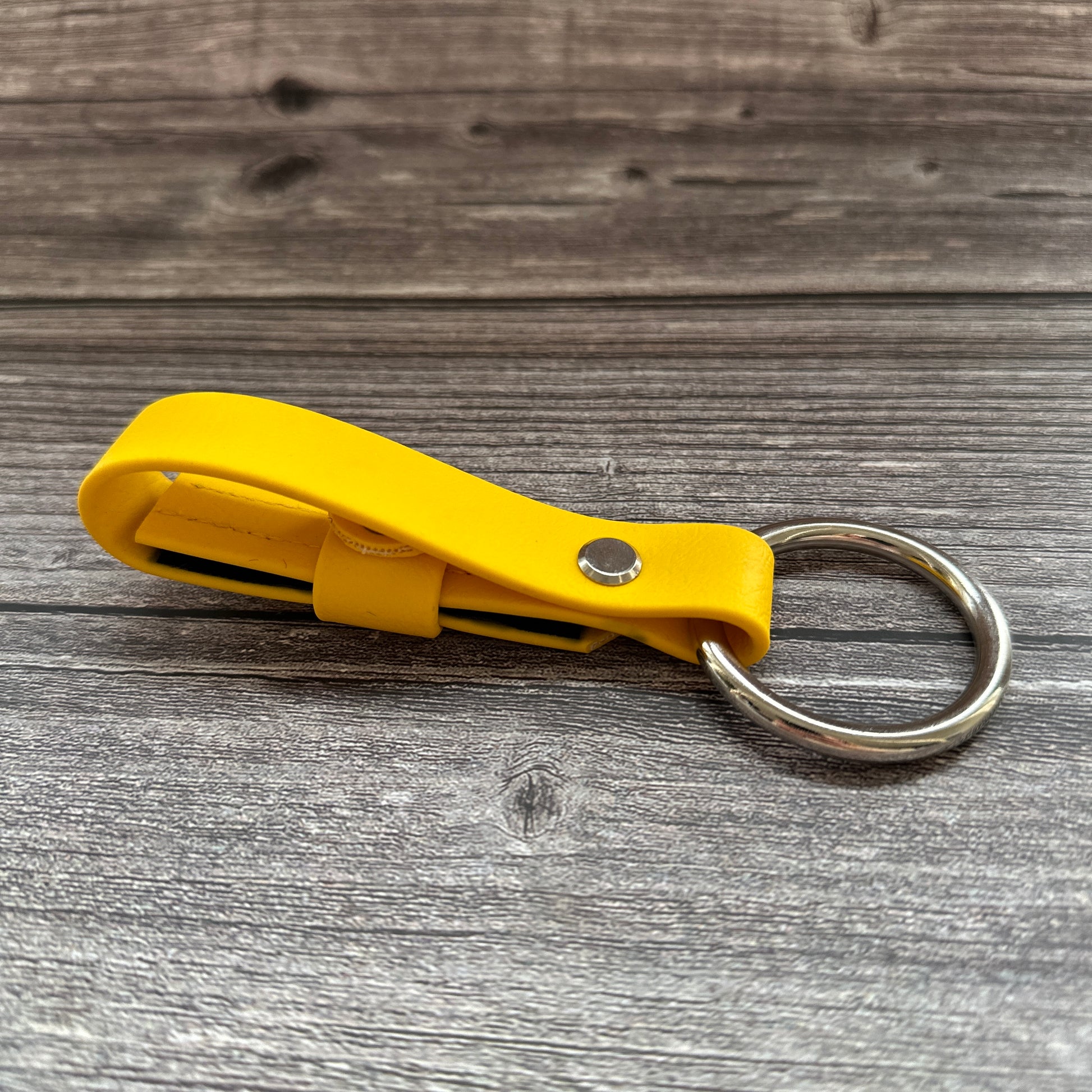 Eine gelbe Leder StallSchätze Sicherheitsschlaufe für Halfter mit einem silbernen Schlüsselring liegt auf einer Holzfläche, gefaltet und mit einer Metallniete gesichert, und bietet eine Option zur Personalisierung.