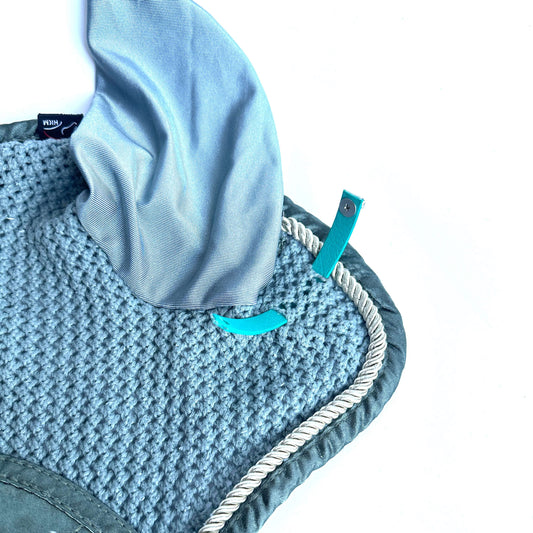 Eine Nahaufnahme der StallSchätze Fliegenohren-Clips (2 Stück) zeigt eine strukturierte blaue Polsterung mit gewellten Rändern, cremefarbenen Seilbesatz und zwei blaue Riemen mit Druckknöpfen. Darüber ist ein glattes, hellblaues Tuch vor weißem Hintergrund drapiert.