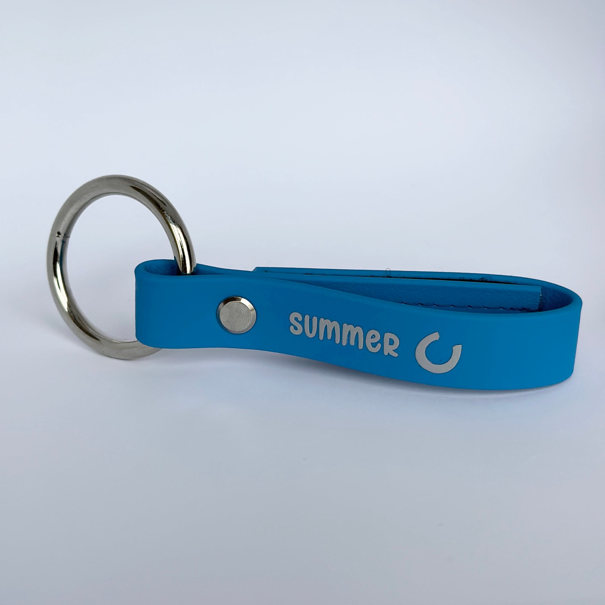 Die StallSchätze Sicherheitsschlaufe für Halfter ist ein blauer Schlüsselanhänger mit einem Metallring, auf dem "SUMMER" und ein kreisförmiges Pfeilsymbol in weißer Farbe auf einem schlichten weißen Hintergrund abgebildet sind.
