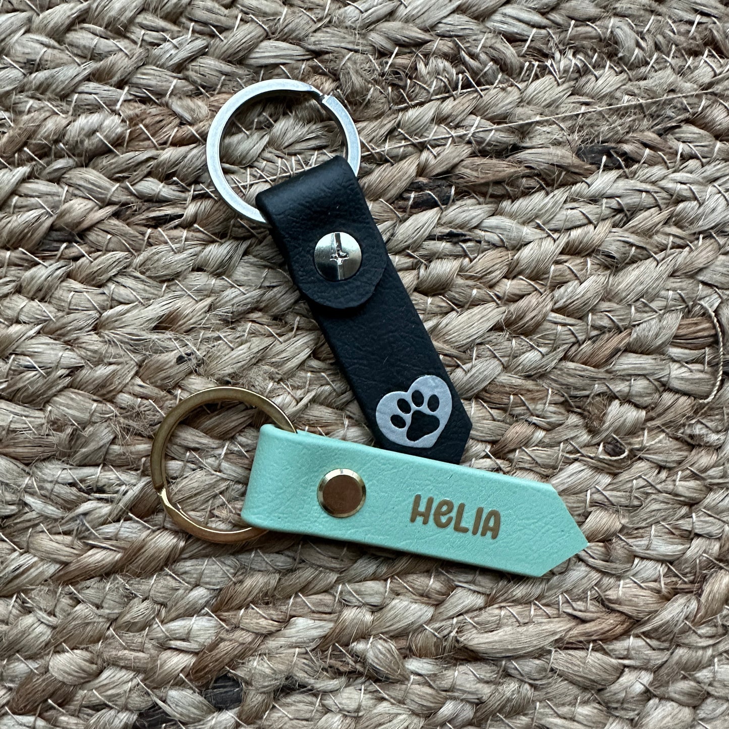 Zwei StallSchätze aus Leder auf einer gewebten Matte: der eine schwarz mit silbernem Pfotenabdruck im Hundemarken-Stil, der andere mintgrün mit goldenen Beschlägen und HELIA in goldenen Buchstaben, perfekt passend zu jedem schicken Halsband.