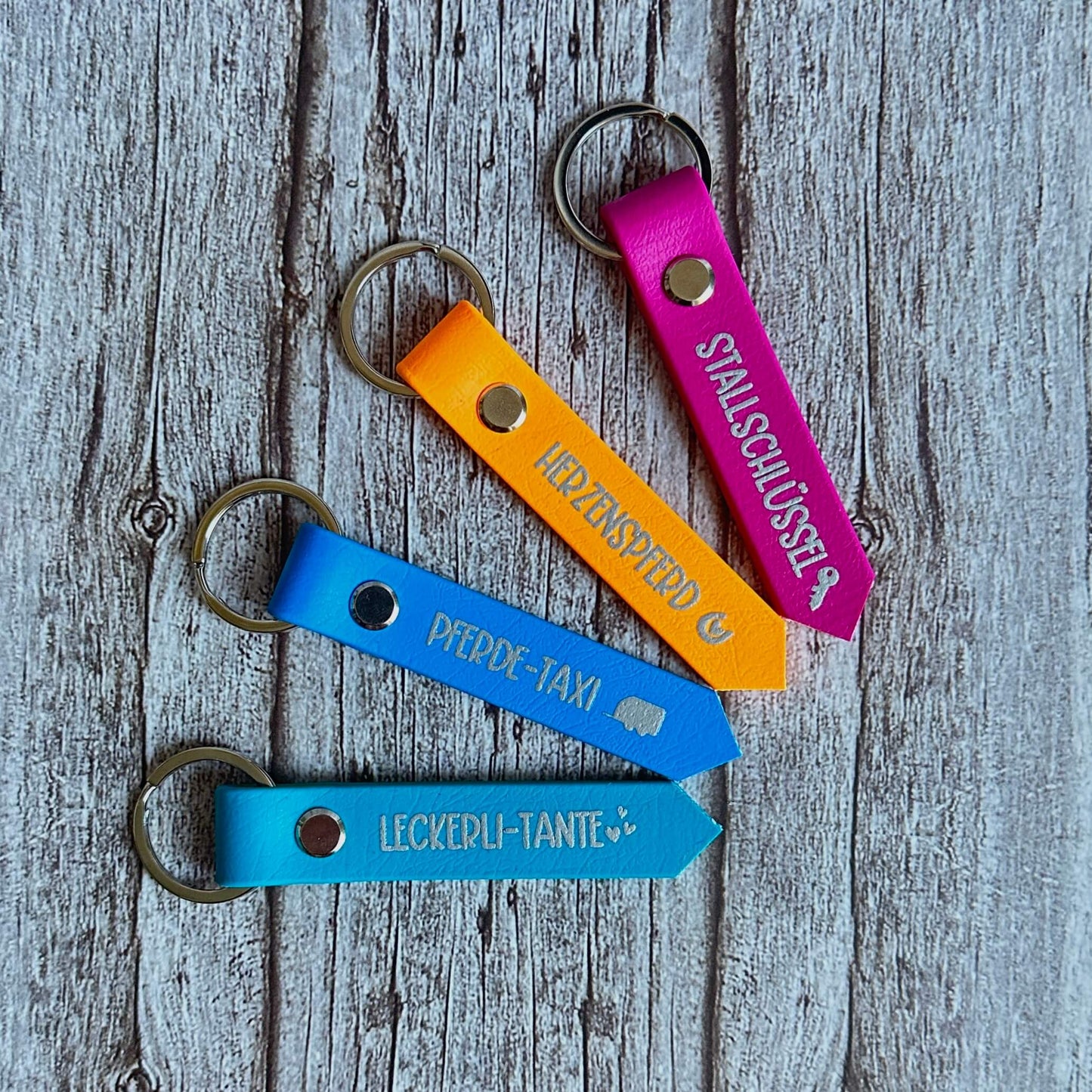 Vier farbenfrohe Biothane-Schlüsselanhänger von StallSchätze - rosa, orange und zwei blaue - zeigen jeweils deutsche Wörter wie Pferde-Taxi" oder Stallschlüssel" mit Piktogrammen, die diagonal auf einer rustikalen Holzoberfläche angeordnet sind.
