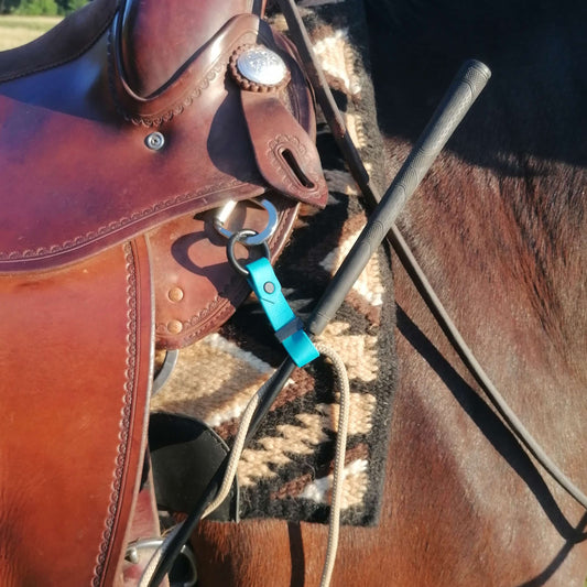 Nahaufnahme eines braunen Ledersattels auf einem Pferd, mit dem StallSchätze Gertenhalter für Spring- und Dressurgerten in Blau an einem Ring befestigt, mit einer schwarzen Gerte und einer gemusterten Satteldecke darunter.