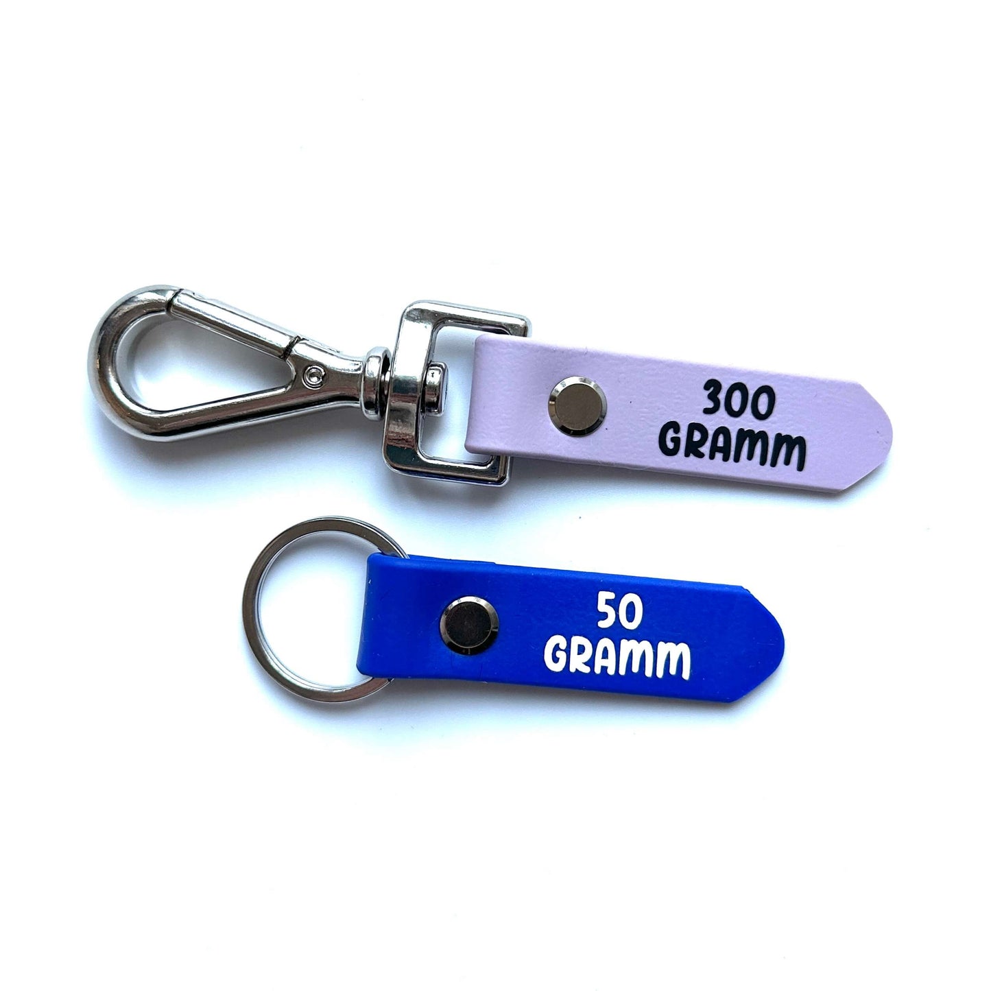Abgebildet sind zwei Schlüsselanhänger: ein lila *NEU* Gramm-Anhänger Beschriftung für Winterdecken mit Metallschließe (300 GRAMM) und ein blauer mit Schlüsselring (50 GRAMM), beide von StallSchätze. Ideale Ergänzung für Ihre StallSchätze-Sammlung!.