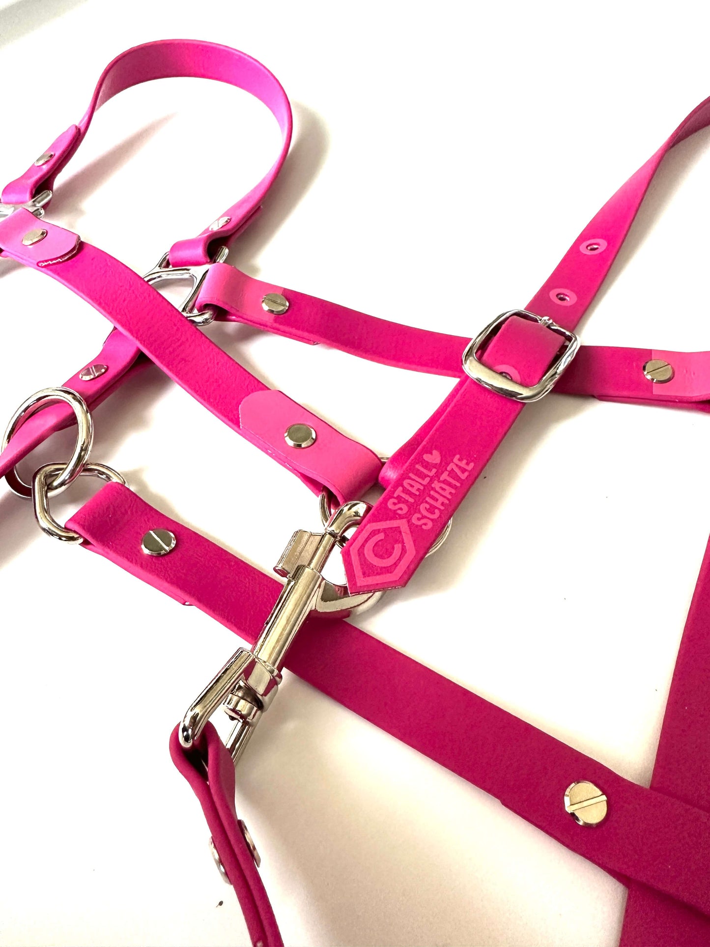 Ein leuchtend rosa, verstellbares Biothane-Halfter mit hochwertigen Details von StallSchätze liegt auf weißem Untergrund. Ein kleines pinkes Etikett mit Logo bietet Personalisierungsmöglichkeiten nahe der silbernen Schnalle.