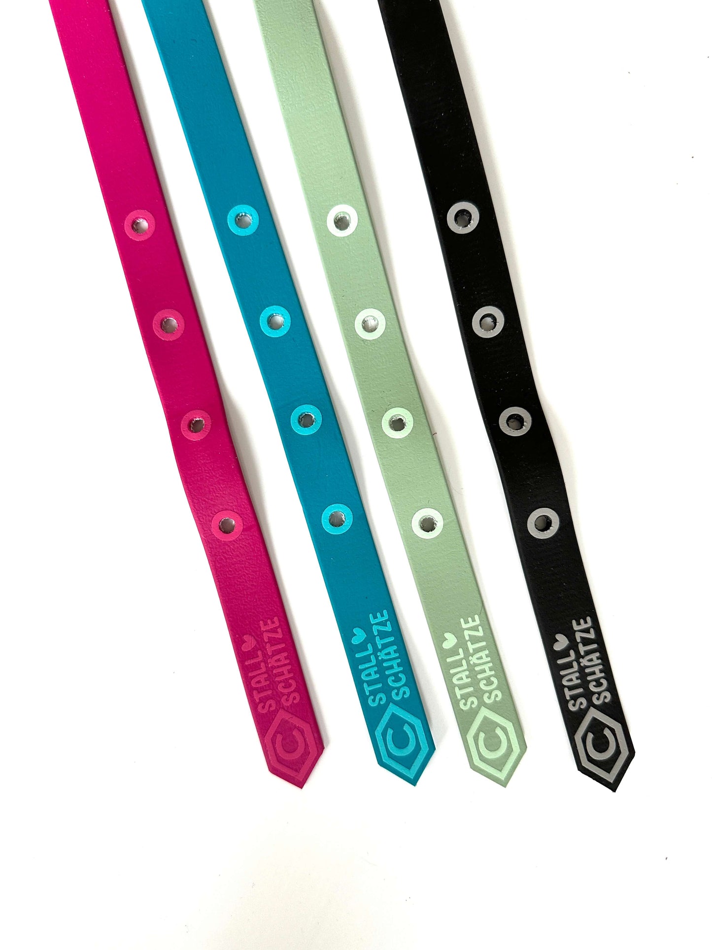 Vier bunte, verstellbare Biothane-Halfter-Armbänder von StallSchätze (rosa, blau, grün, schwarz) mit Metallösen sind diagonal angeordnet; die grünen und schwarzen Bänder zeigen ein Herz und "STALL SCHATZ" in weiß.