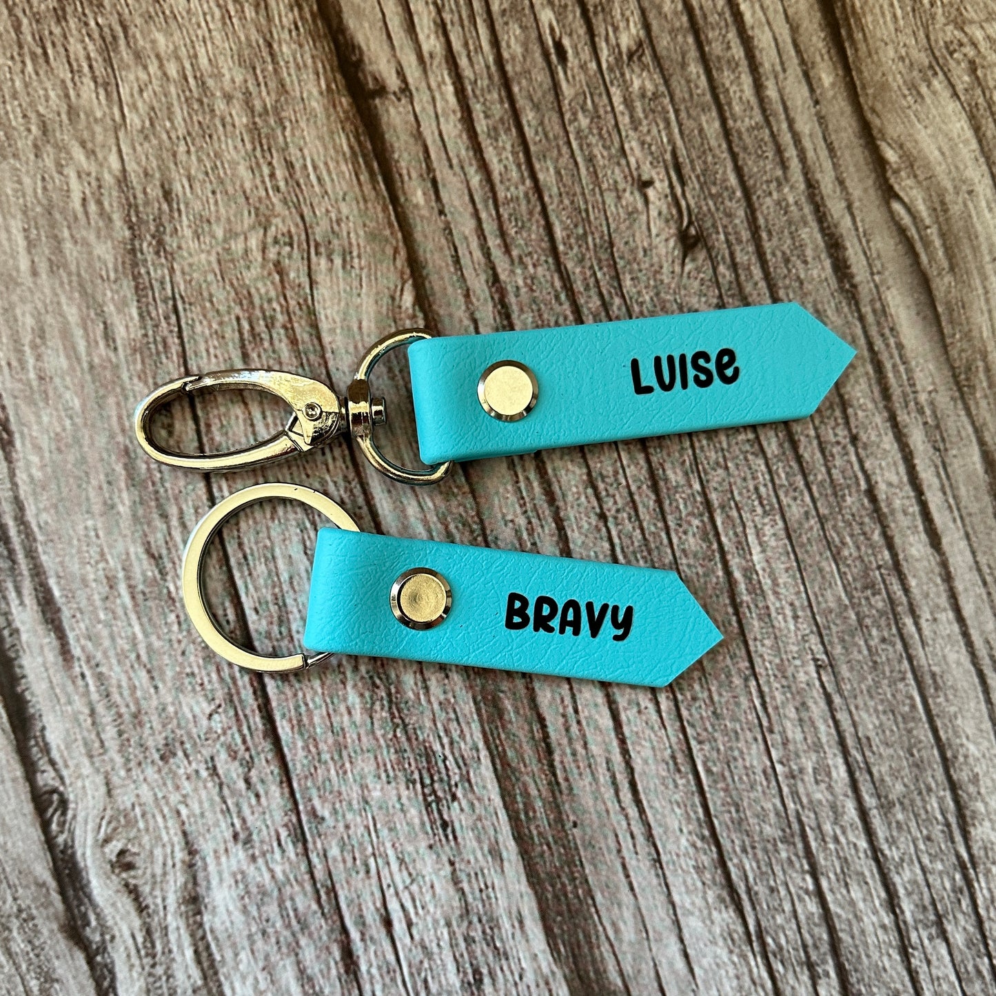 Zwei blaue Leder SOS-Notfallmarken für Sattel und Halfter von StallSchätze mit goldenen Druckknöpfen und Metallringen liegen auf Holz. Jeweils personalisiert mit LUISE oder BRAVY-ideal für Pferdenamen oder eine Notfallnummer.