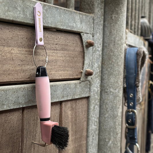 Ein rosafarbener StallSchätze MagBarn Magnet-Anhänger für Hufkratzer & Co hängt an einem Schlüsselring an einer Stalltür aus Metall und Holz, daneben ein blaues Pferdehalfter als weiterer praktischer Stallhelfer.
