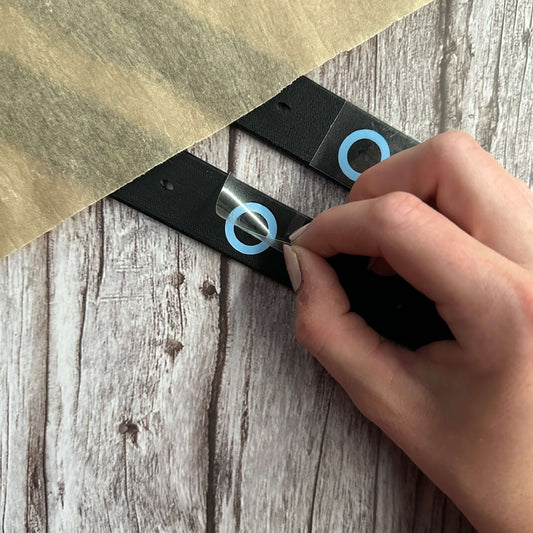 Eine Hand zieht durchsichtiges Klebeband von einem schwarzen Steigbügelriemen-Längen-Kennzeichner von StallSchätze mit blauen Kreise und Löcher ab, der neben einem Blatt Pergamentpapier auf einem Holztisch liegt.