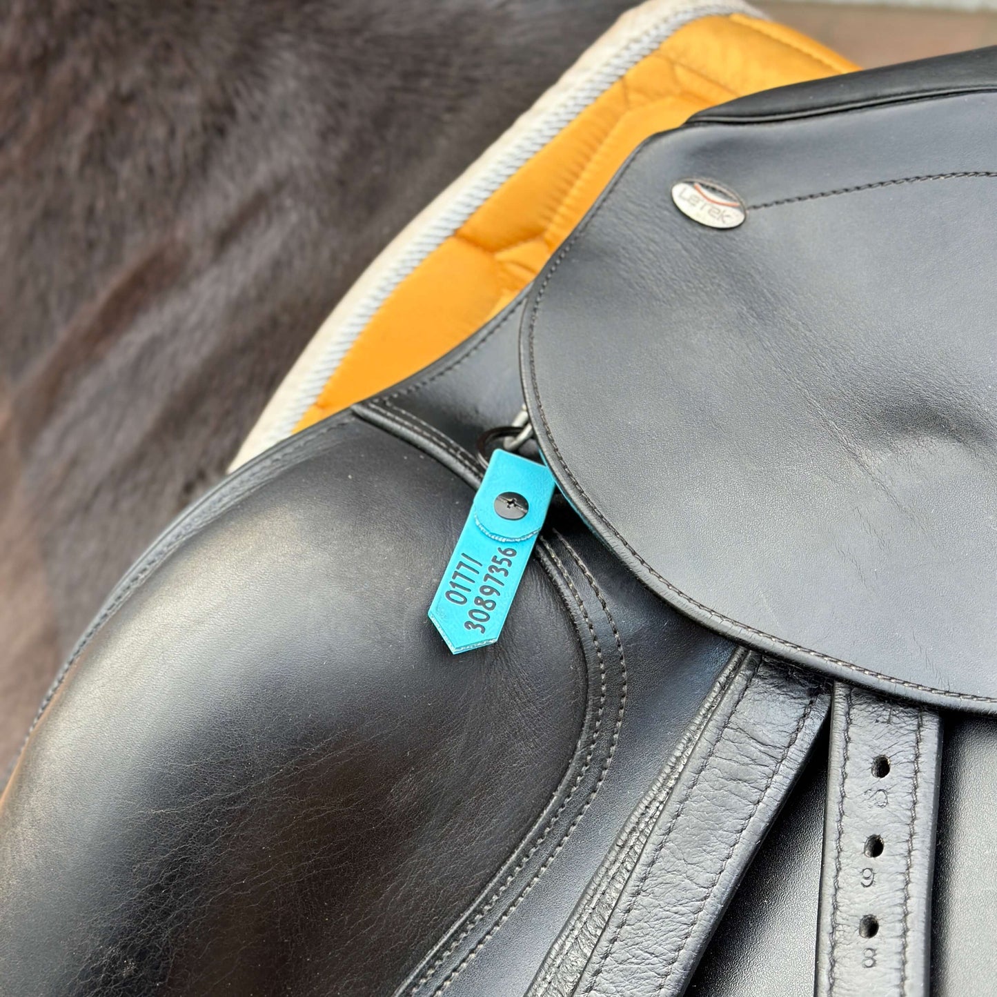 Nahaufnahme eines schwarzen Ledersattels auf einem Pferd mit einer angebrachten StallSchätze SOS-Notfallmarke für Sattel und Halfter, beschriftet mit OTTI 30897356. Darunter ist eine gelbe Schabracke zu sehen.
