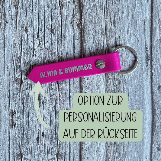 Ein rosafarbener Biothane-Schlüsselanhänger von StallSchätze mit "ALINA & SUMMER" in weißer Schrift liegt auf einer Holzfläche. Ein Pfeil weist auf die Möglichkeit hin, die Rückseite zu personalisieren. Ideal als Stallschlüssel!.