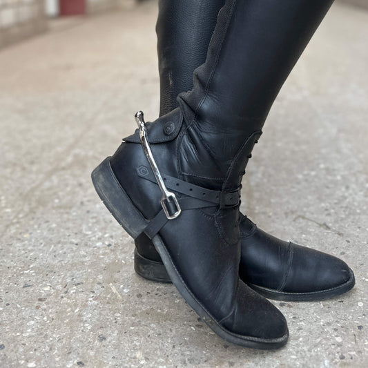 Nahaufnahme einer Person in schwarzen Lederreitstiefeln mit StallSchätze Sporenriemen und schwarzer Hose, stehend auf Beton. An einem Stiefel ist ein Steigbügel aus Metall befestigt.