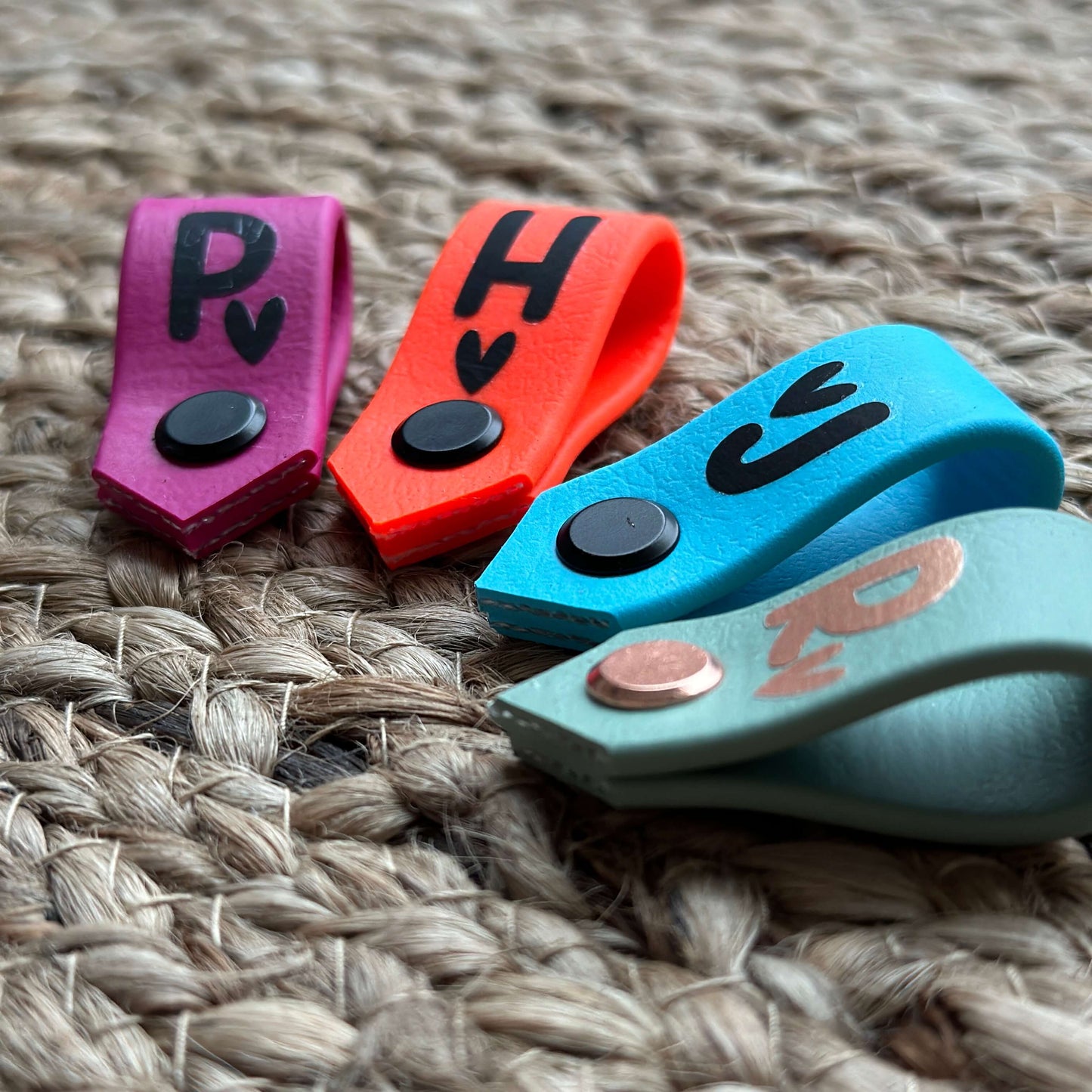 Vier Mini Halfter-Tags auf einem Bast-Hintergrund in den Farben beere, neon-orange, cyan und salbei, bedruckt mit Buchstaben.