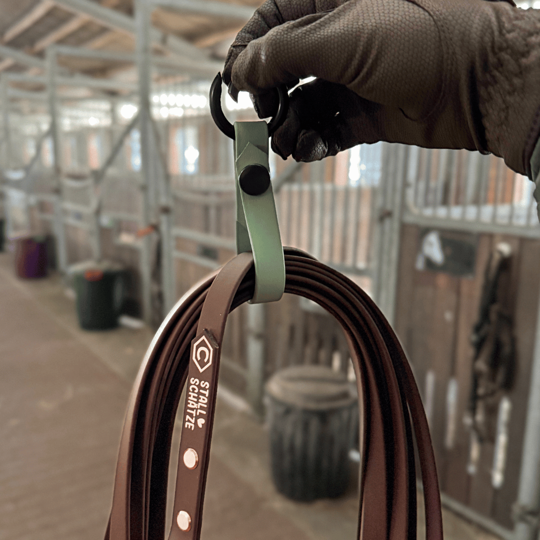 Eine behandschuhte Hand hält die StallSchlaufe - der praktische Longenhalter von StallSchätze, einen grünen Haken mit schwarzer Schließe zum Aufhängen von braunen Lederzügeln, Doppellongen oder Longen in einem Stall mit Holzboxen und unscharfem Hintergrund.