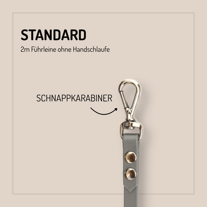 Eine graue StallSchätze Biothane-Hundeleine 2m, individuell konfigurierbar, mit Metallnieten und Karabinerhaken ist senkrecht dargestellt. Der Text lautet STANDARD, 2m Führleine ohne Handschlaufe, SCHNAPPKARABINER mit einem Pfeil auf den Karabinerhaken.
