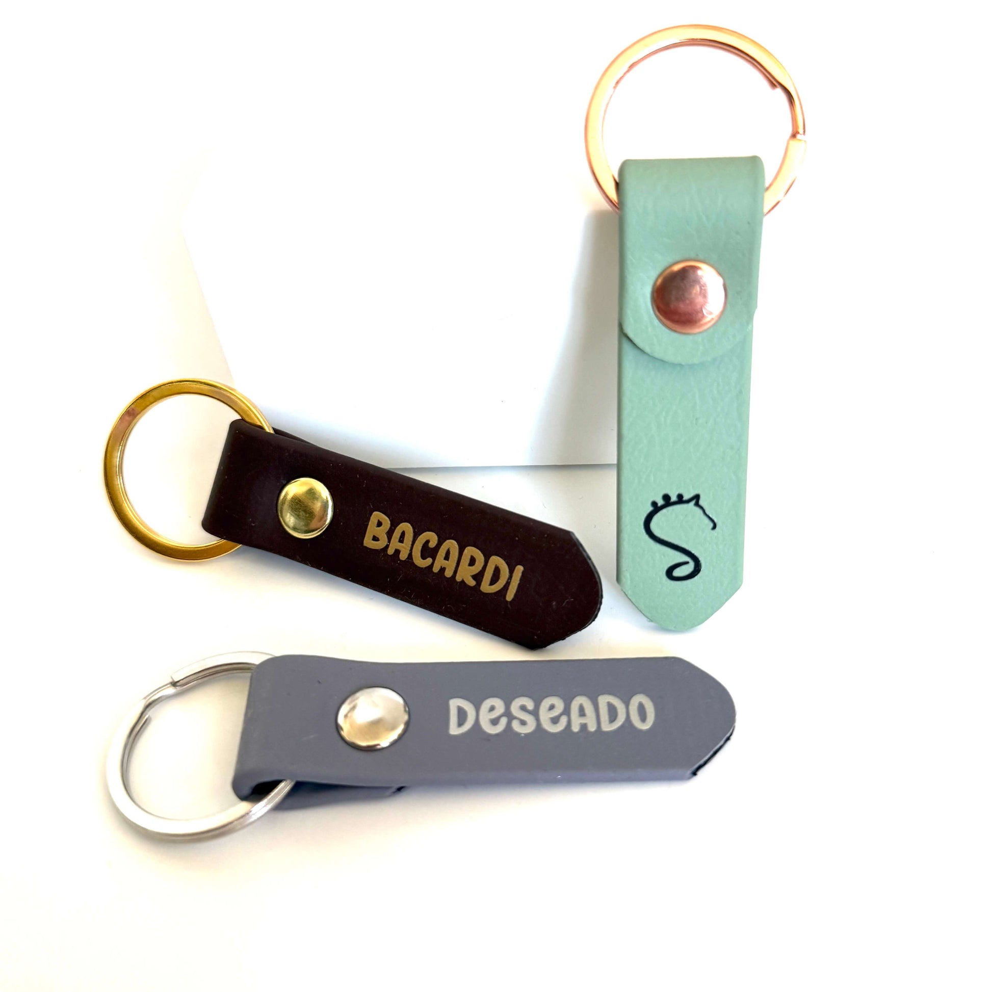 Entdecken Sie drei personalisierte Anhänger-Schlüsselanhänger mit Halfter- und Strick-Beschriftung von StallSchätze aus stilvollem Leder: braun („BACARDI“), grün mit einem Seepferdchen-Symbol, das an ein Glückshufeisen erinnert, und grau („DESEADO“). Jeder verfügt über einen farblich abgesetzten Metall-Schlüsselring, was sie zu einzigartigen und modischen Halfter-Anhängern für Ihre Schlüssel macht.
