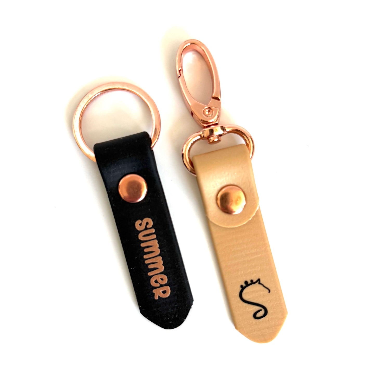 Zwei Leder-Schlüsselanhänger mit roségoldenen Beschlägen – ein schwarzer mit dem weißen Aufdruck „summer“ und ein beiger mit einem kleinen dekorativen „S“, einer Kronenillustration und einem dezenten Glückshufeisen-Detail – sind vor weißem Hintergrund abgebildet. Diese „Personalisierten Anhänger mit Halfter- & Strick-Beschriftung“ von StallSchätze sind stilvolle Anhänger mit Pferdenamen für Pferdeliebhaber.