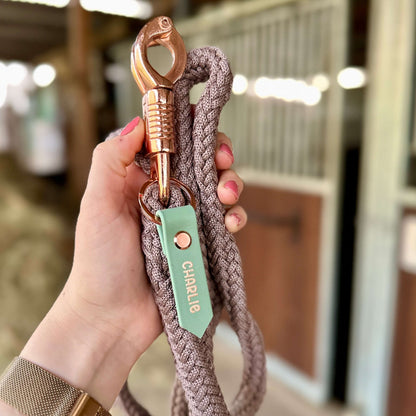 Eine Hand hält eine geflochtene Seil-Leine mit einem glänzenden Messing-Clip und einer mintgrünen StallSchätze Personalisierter Anhänger Halfter- & Strick-Beschriftung mit der Aufschrift CHARLIE. Im Hintergrund sind unscharfe Stalltüren zu sehen.