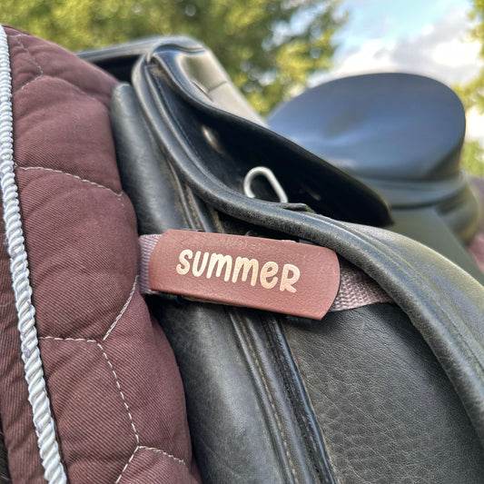 Eine Nahaufnahme zeigt die StallSchätze Schabracken-Beschriftung auf einem braunen, gesteppten Pad und einem schwarzen Ledersattel. Ein weißes Band mit der Aufschrift SUMMER hebt sich ab, mit verschwommenen grünen Bäumen und blauem Himmel im Hintergrund.