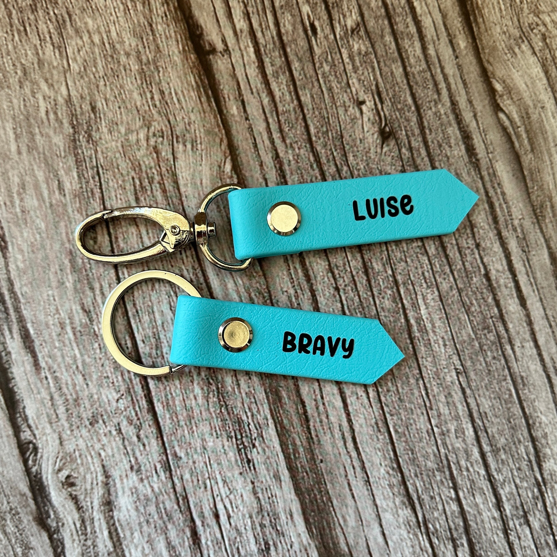 Zwei blaue Leder SOS-Notfallmarken für Sattel und Halfter von StallSchätze mit goldenen Druckknöpfen und Metallringen liegen auf Holz. Jeweils personalisiert mit LUISE oder BRAVY-ideal für Pferdenamen oder eine Notfallnummer.