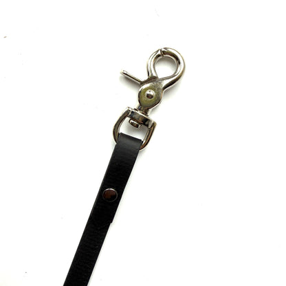 Eine Nahaufnahme der StallSchätze Safety-Schweifriemen mit Sollbruch-Stelle aus schwarzem Biothane mit silbernem Drehverschluss, dargestellt vor einem weißen Hintergrund.