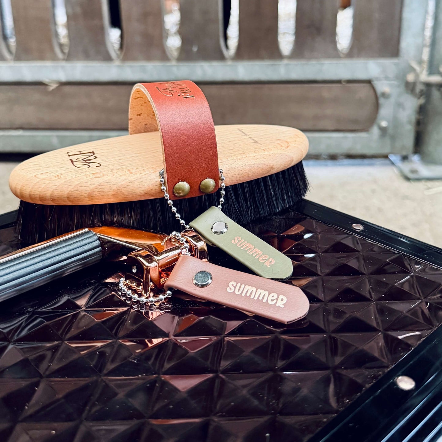 Eine hölzerne Pflegebürste mit einem braunen Ledergriff und zwei StallSchätze Anhänger zum Beschriften von Bürsten und Putzkästen mit der Aufschrift "Sommer" liegen auf einer strukturierten schwarzen Oberfläche neben einem Metallgitter.