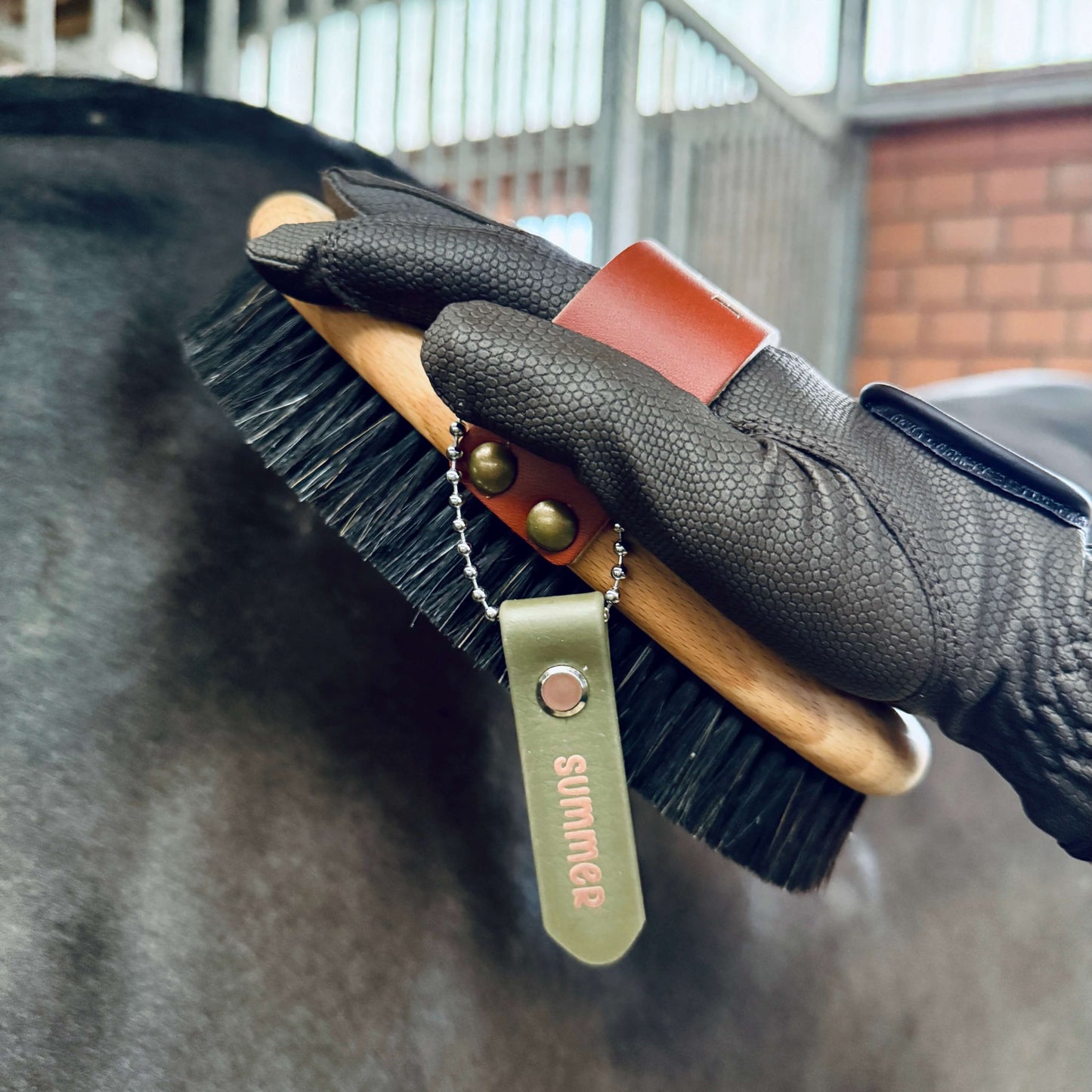 Eine behandschuhte Hand bürstet ein Pferd mit einer hölzernen Bürste, die mit dem "Anhänger zum Beschriften von Bürsten und Putzkästen" von StallSchätze beschriftet ist, aus einem Pflegeset in einem Stall mit Metallstangen und Ziegelwänden.