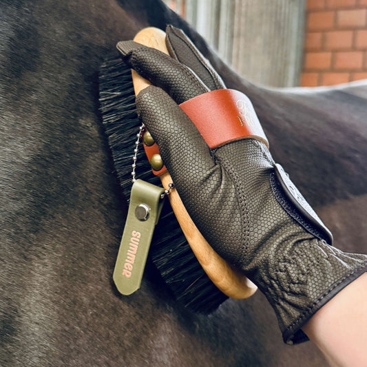 Eine behandschuhte Hand bürstet das dunkle Fell eines Pferdes mit einer Bürste, die mit dem StallSchätze Anhänger zum Beschriften von Bürsten und Putzkästen beschriftet ist; im unscharfen Hintergrund sind eine Stallwand, Metallstangen und ein Teil eines Putzkastens zu sehen.