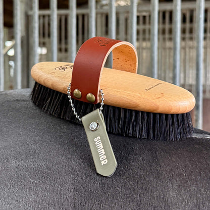 Eine hölzerne Kardätsche mit schwarzen Borsten und einem braunen Lederriemen ruht auf dem Rücken eines Pferdes. An dem Riemen hängt ein StallSchätze Anhänger zum Beschriften von Bürsten und Putzkästen mit der Aufschrift "Sommer"; andere Bürsten sind im Hintergrund verschwommen.