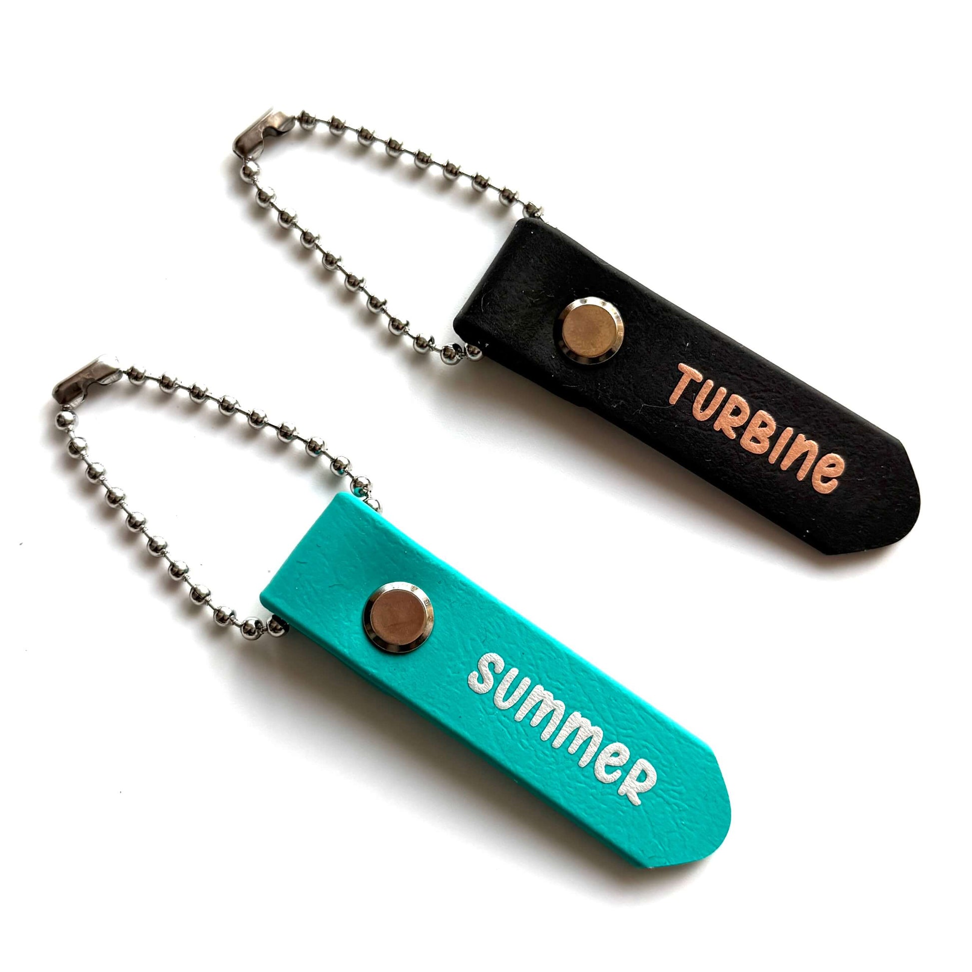 Zwei StallSchätze "Anhänger zum Beschriften von Bürsten und Putzkästen" als Schlüsselanhänger: einmal schwarz mit TURBINE (pfirsichfarbene Buchstaben), einmal teal mit SUMMER (weiß). Beide haben Metallketten und Druckknöpfe - ideal für Putzkasten oder Bürsten.