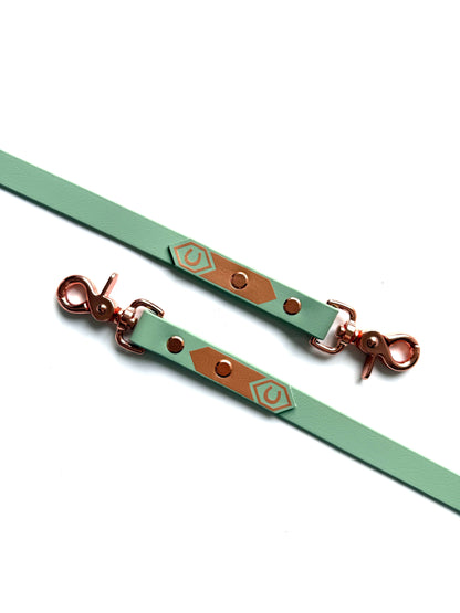 Ein mintgrüner StallSchätze Biothane-Zügel mit Scherenkarabinern, mit roségoldenen Clips und braunen geometrischen Akzenten auf weißem Hintergrund. Reißfest und leicht zu reinigen-ideal für den täglichen Gebrauch.