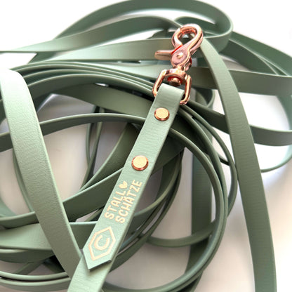 Eine Nahaufnahme der salbeigrünen StallSchätze Biothane-Longe 8 Meter Leine mit roségoldenen Beschlägen, goldgeprägtem Etikett und Hexagon-Logo. Diese pflegeleichte Ausrüstung ist locker auf eine weiße Oberfläche gewickelt.