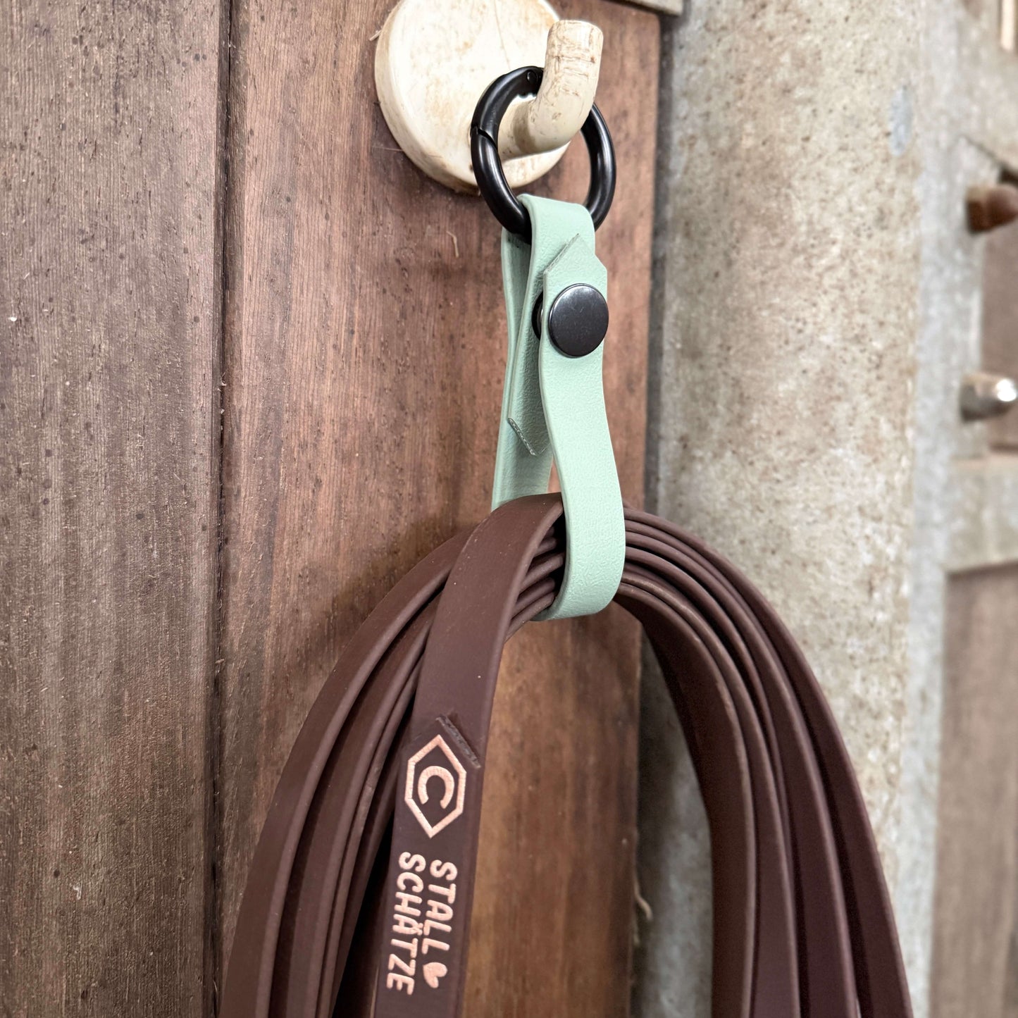 An einem Haken an einer Holzwand hängt eine braune StallSchlaufe - der praktische Longenhalter von StallSchätze mit einem mintgrünen Band, das mit dem StallSchätze-Logo und -Text versehen ist.
