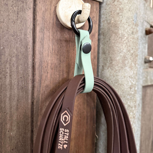 An einem Haken an einer Holzwand hängt eine braune StallSchlaufe - der praktische Longenhalter von StallSchätze mit einem mintgrünen Band, das mit dem StallSchätze-Logo und -Text versehen ist.