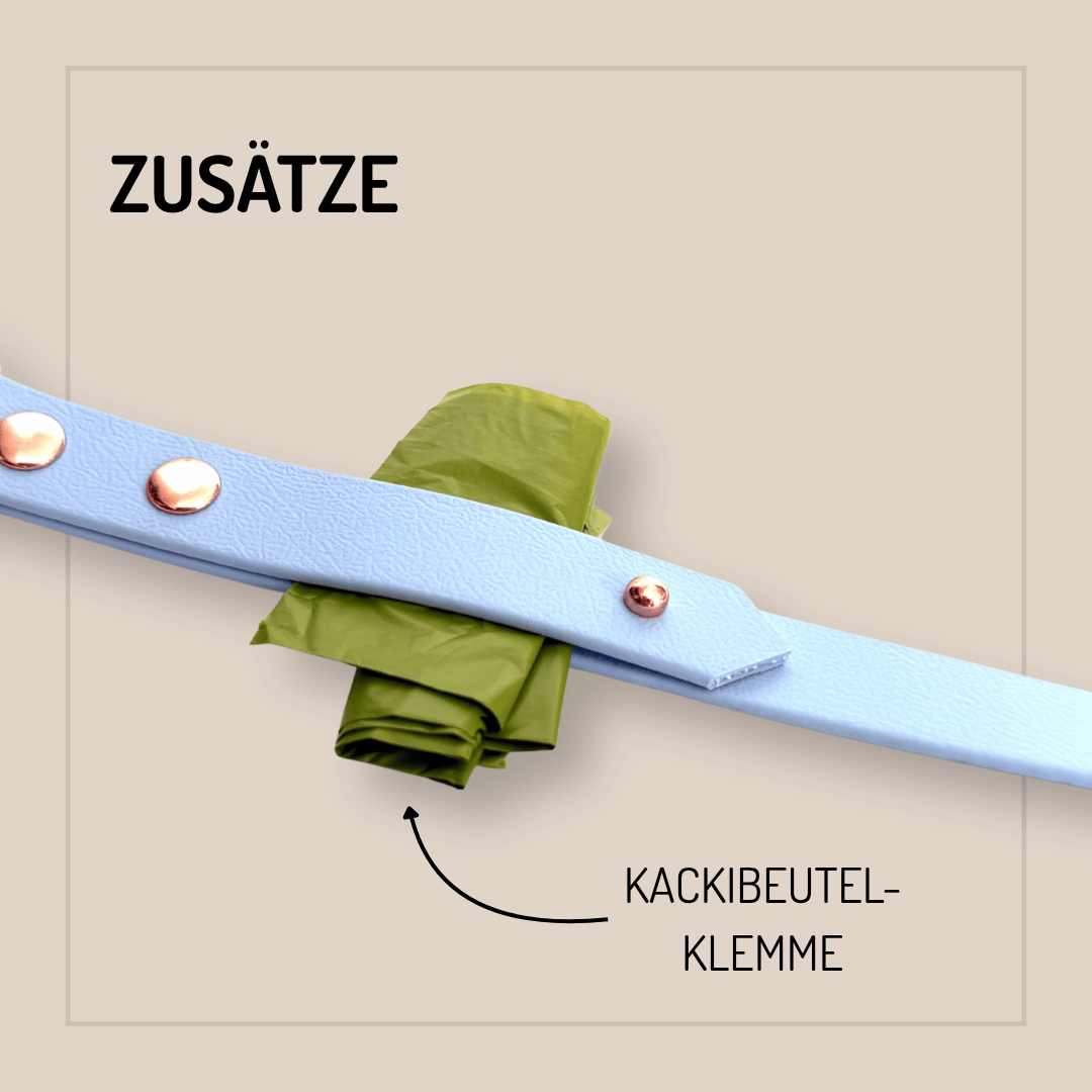 Ein hellblaues Band mit Metallnieten hält grüne Müllbeutel, ideal als Zubehör für Ihre StallSchätze Biothane-Hundeleine 2m - individuell konfigurierbar. Auf dem Etikett steht ZUSÄTZE und KACKIBEUTEL-KLEMME mit einem Pfeil zu den Säcken.