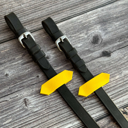 Zwei schwarze Zügelenden, an denen jeweils ein gelber Zügelstopper angebracht ist auf holzigem Untergrund.